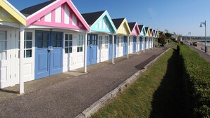 A row of colorful beach huts with white doors and windows, a gravel path, and a grassy area beside a beach. - Corner Barn (Blandford Forum)