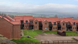 A row of brick buildings with red-tiled roofs, a grassy courtyard, and a view of distant fields and hills.