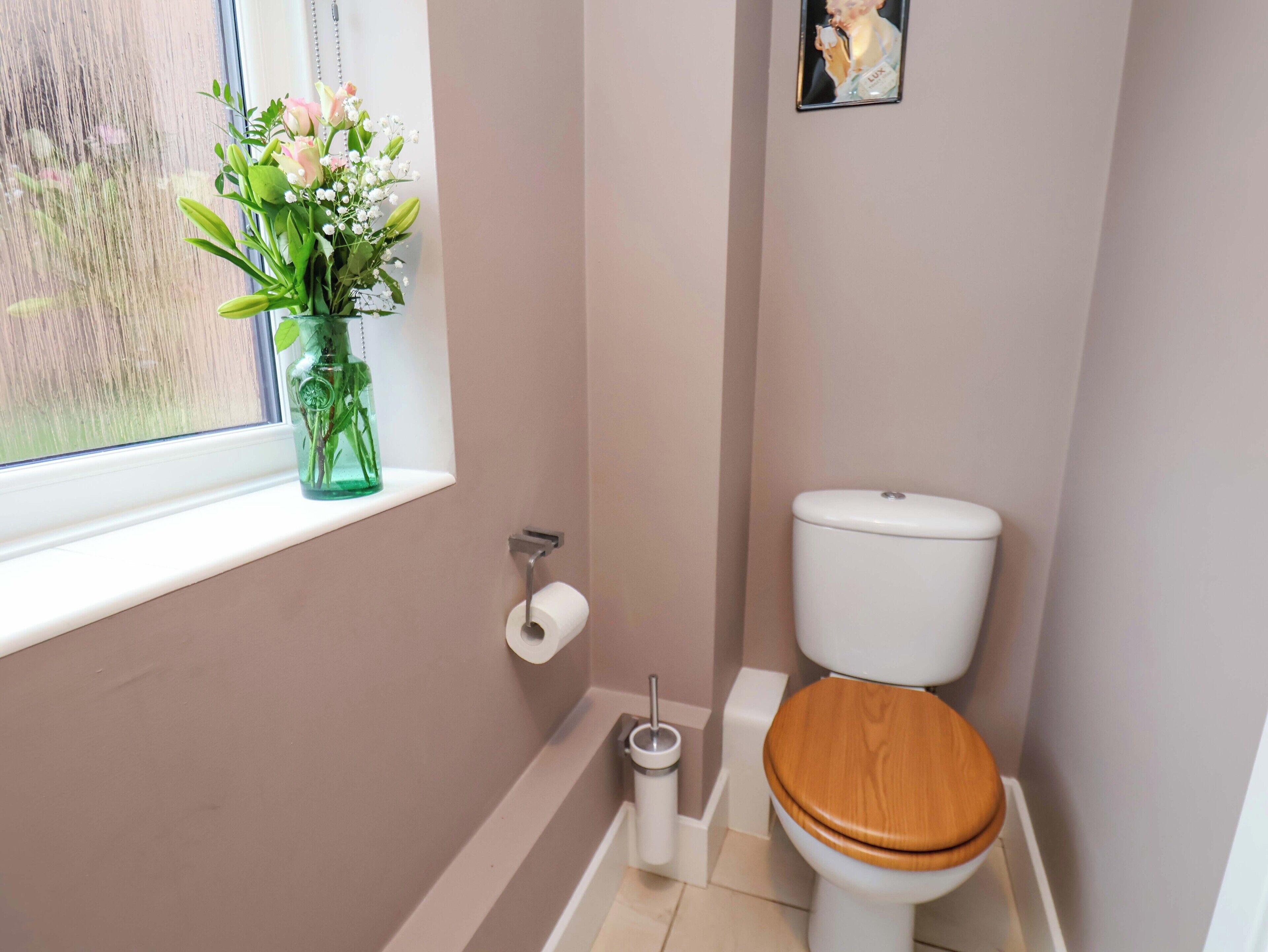 A bathroom with a toilet, a window with a vase of flowers, and a toilet paper holder.