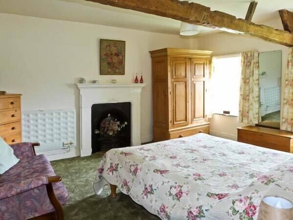 A bedroom with a floral bedspread, a wooden wardrobe, a dresser, a chair, and a fireplace. - High Sprintgill Cottage (Kirkby Stephen)