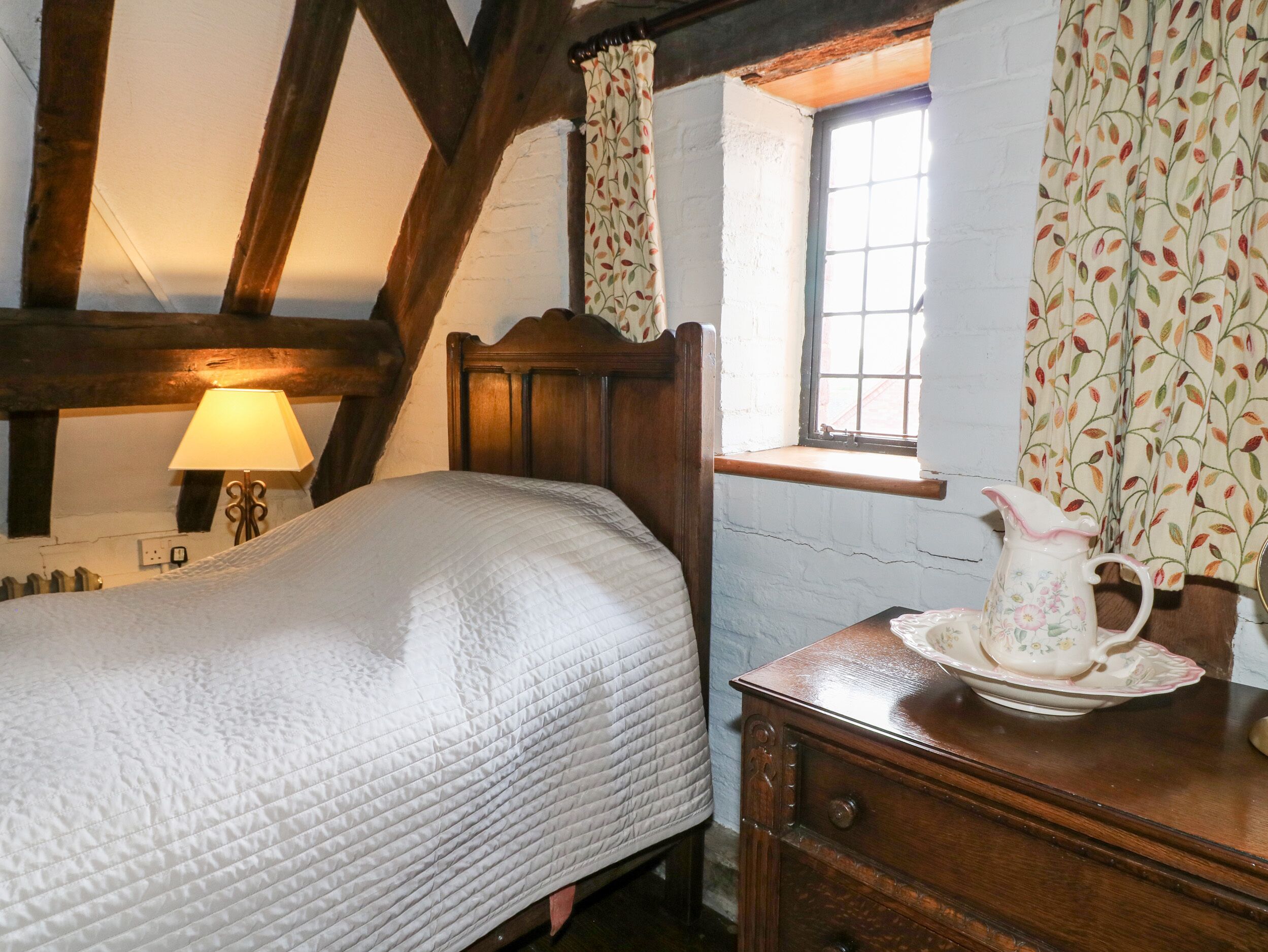 A bedroom with a wooden bed, a bedside table with a pitcher, and a window with curtains.