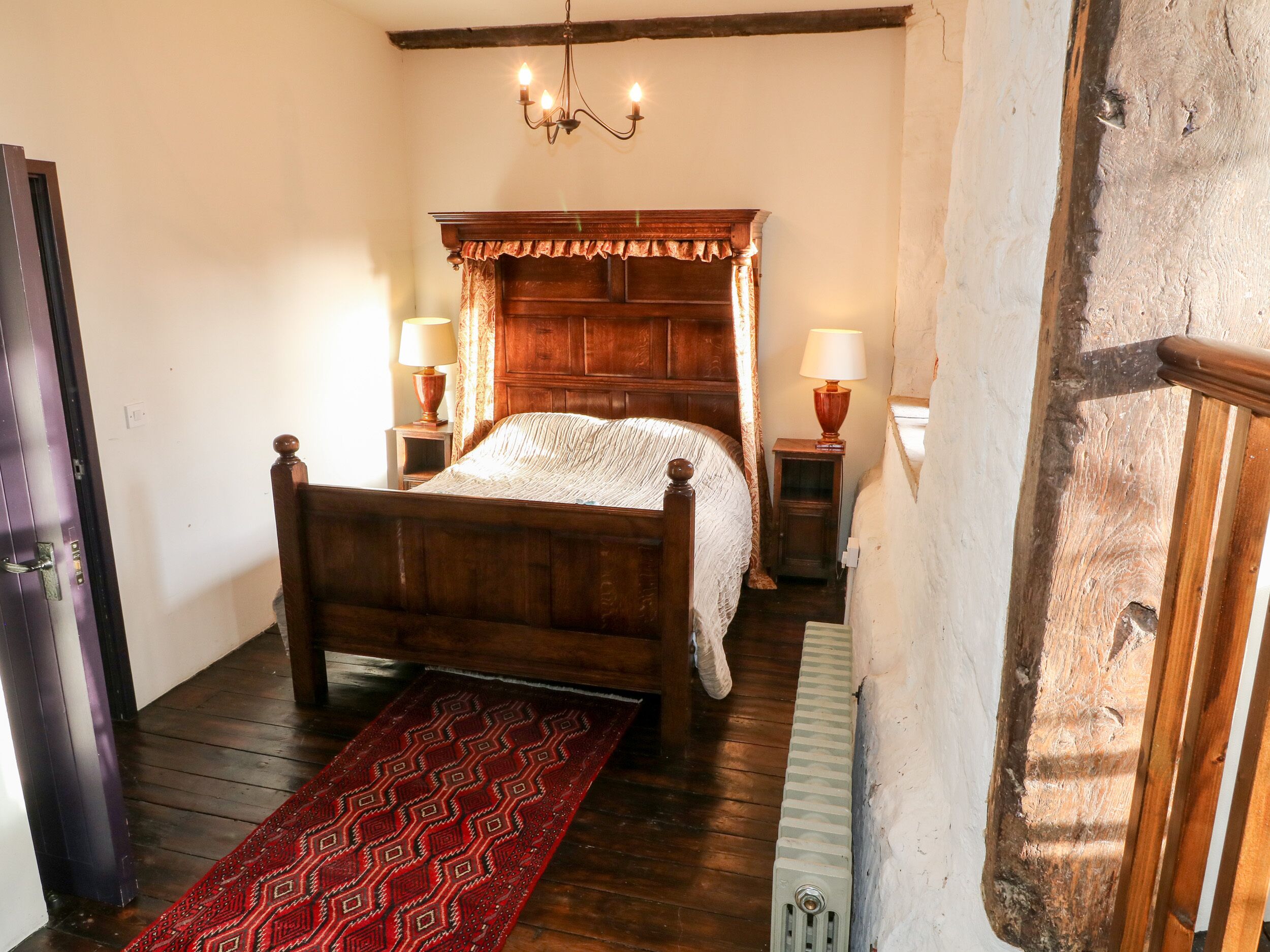 Una cama de madera tradicional con dosel, dos lámparas de noche y una alfombra roja estampada en una habitación con pisos de madera y pared blanca.