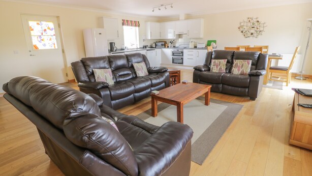 A living room with a brown leather sofa, a wooden coffee table, and a dining area with a wooden table and chairs. - Muir Iosal (Perth)
