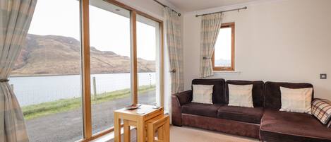 A room with a large window offering a view of a lake and hills, a brown sofa with cushions, and a small wooden table.