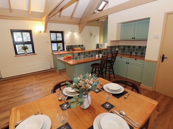 A dining area with a wooden table set for a meal, a kitchen with teal cabinets, and a view of the outdoors through a window. - Foxglove (Fishguard)