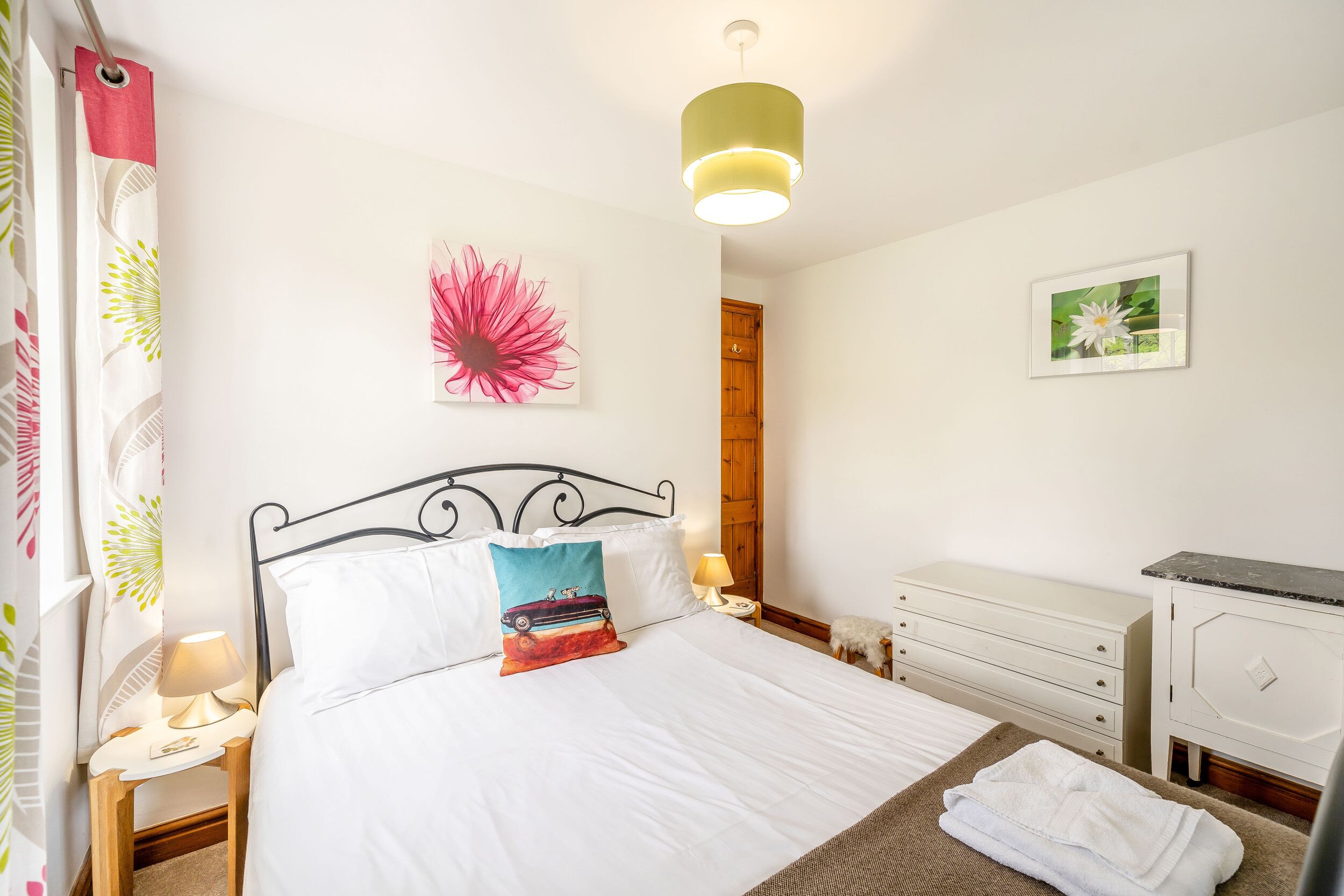 A neatly arranged bedroom with a bed, bedside tables, a lamp, a wooden wardrobe, and a wall with a flower painting.