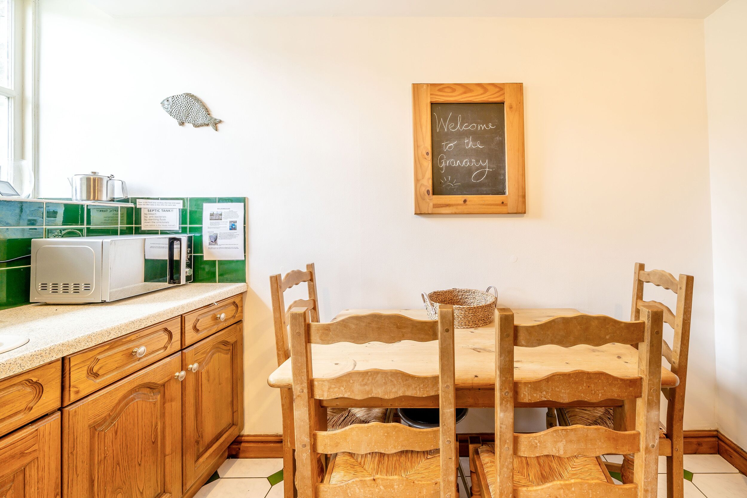 A kitchen with wooden cabinets, a microwave, a chalkboard with a welcome message, and a fish decoration.