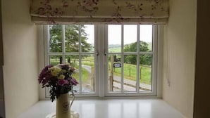 A window with a floral curtain, a vase with flowers, and a view of a rural landscape. - Cockley Beck Cottage (Broughton in Furness)