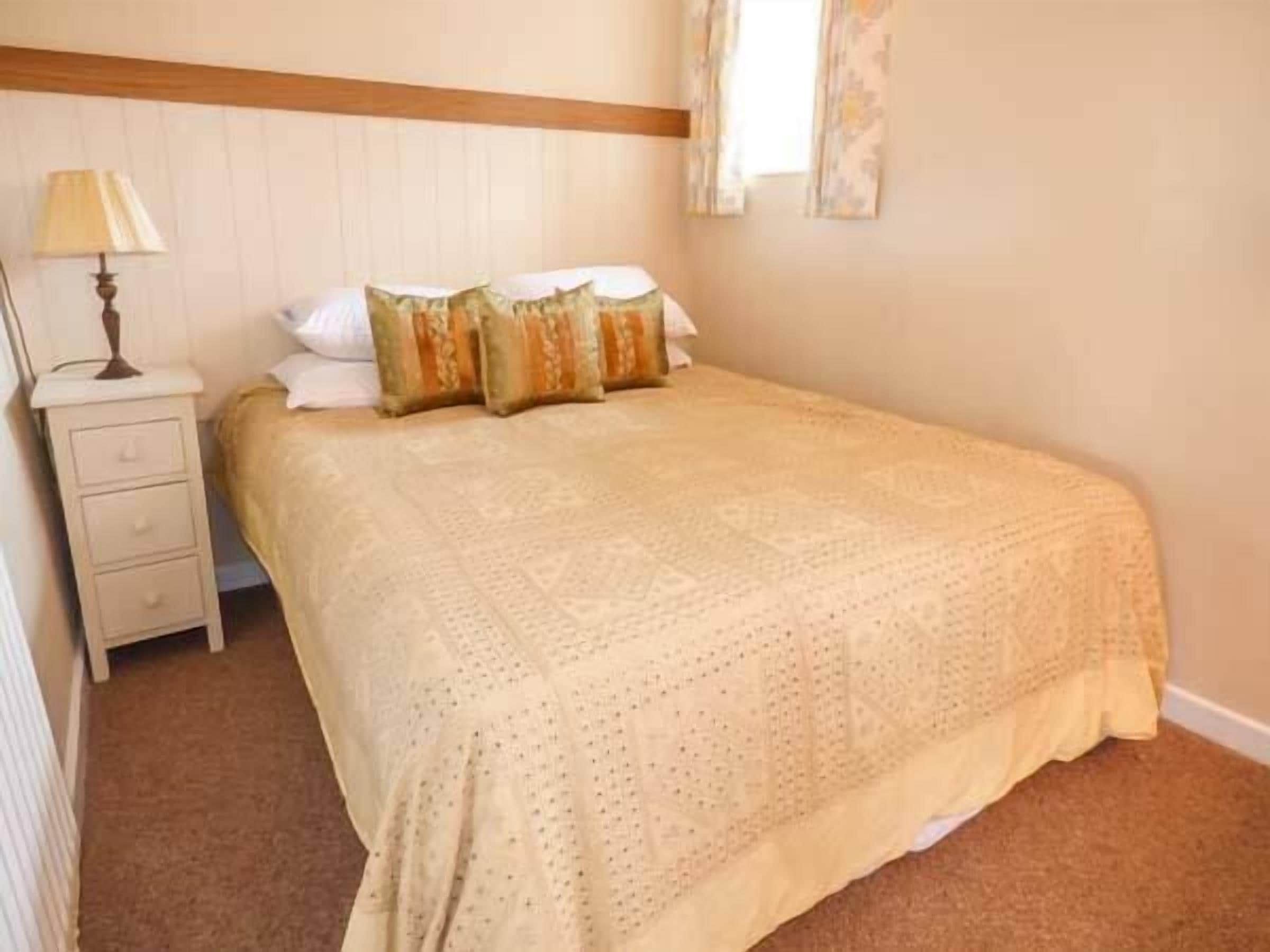 A neatly made bed with a patterned bedspread, a bedside table with a lamp, and a window with curtains.