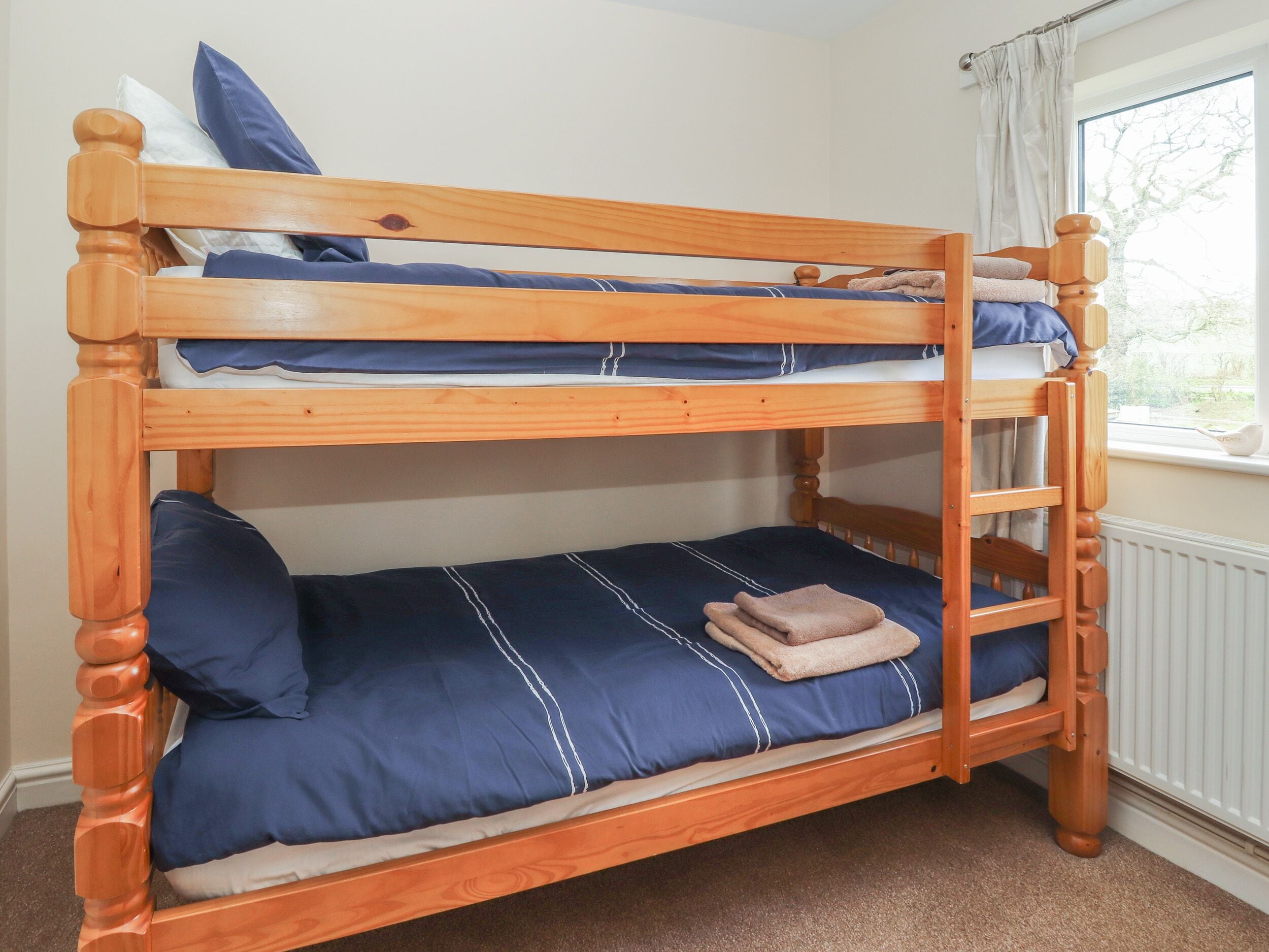 A bunk bed with a wooden frame, blue bedding, and a window with curtains.