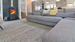 A modern living room with a grey sectional sofa, a large grey rug, a fireplace, and a lofted ceiling with skylights. - Pig House, Boskensoe Barns (Falmouth)