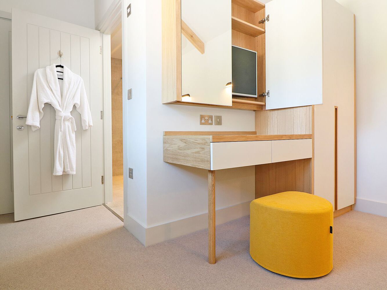 A room with a white robe hanging on a door, a wooden desk with a mirror above it, and a yellow ottoman.