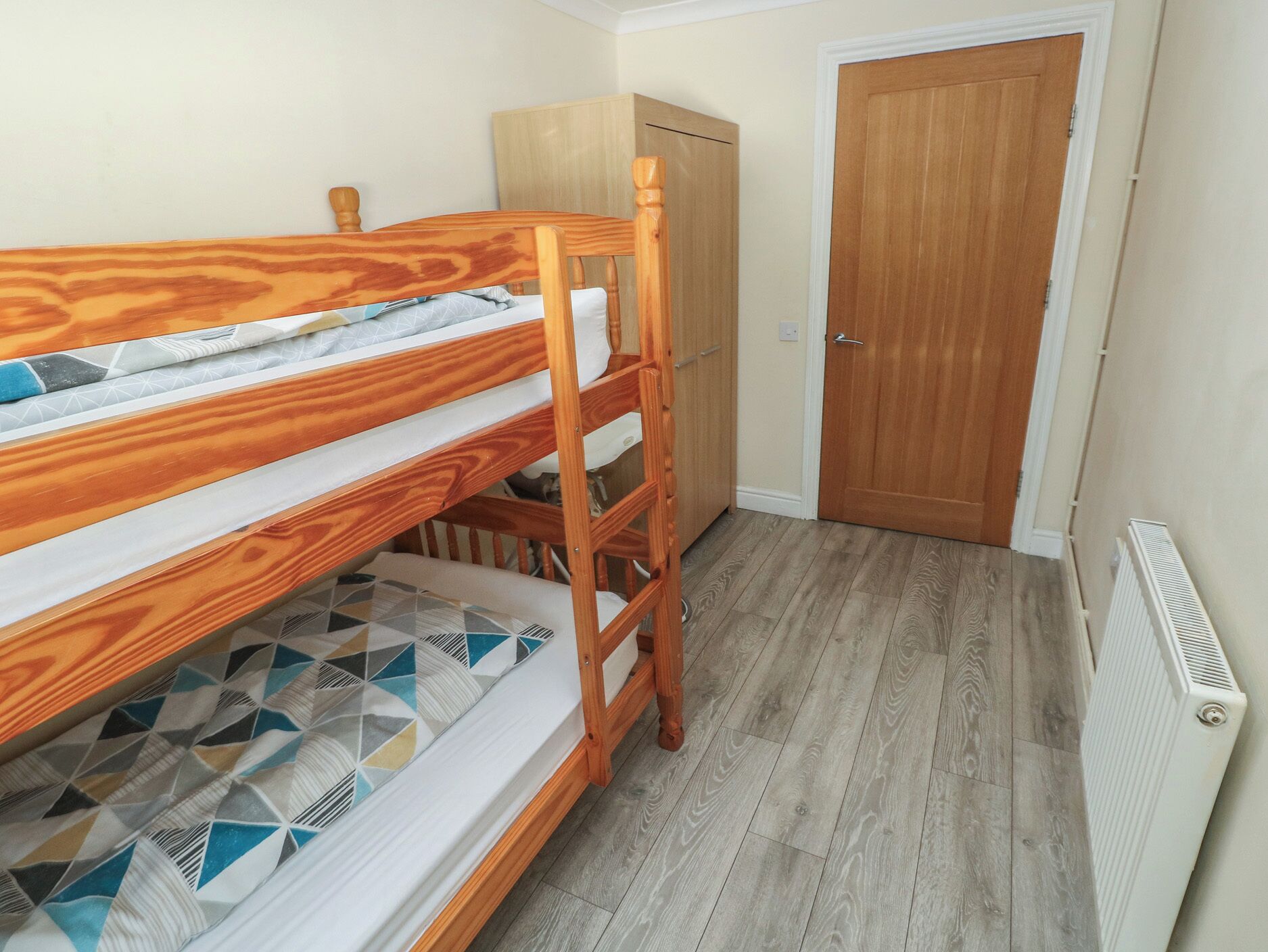 A bunk bed with a geometric-patterned mattress, a wooden wardrobe, and a door in a room with wooden flooring and a radiator.