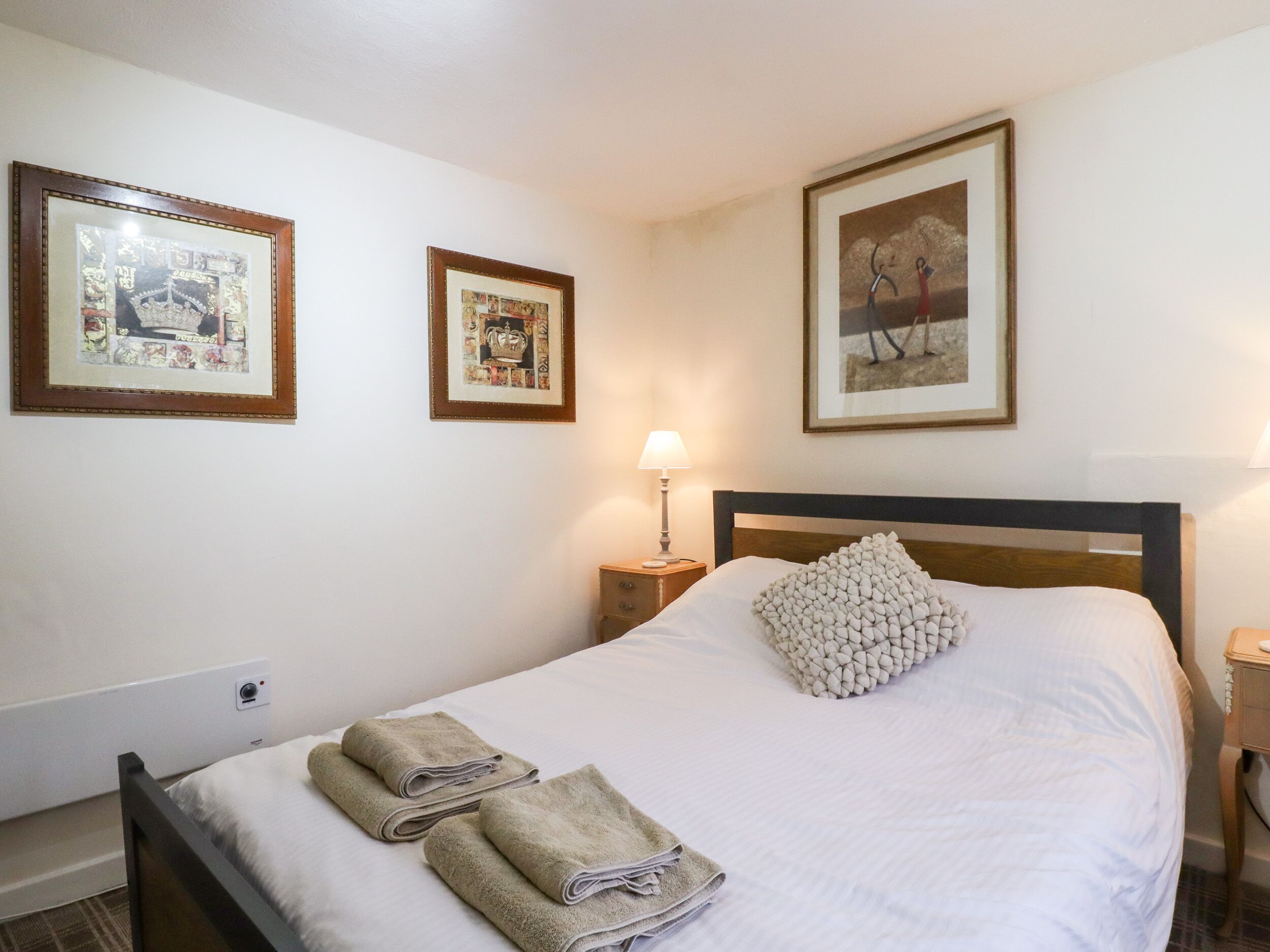 Cama bem arrumada com toalhas, mesa de cabeceira com abajur e quadros nas paredes.