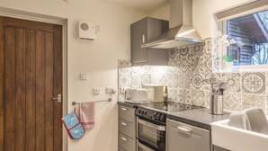 A compact kitchen with a wooden door, a built-in oven, a microwave, and a window with a view of the outside. - Woodpeckers Cottage (Church Stretton)