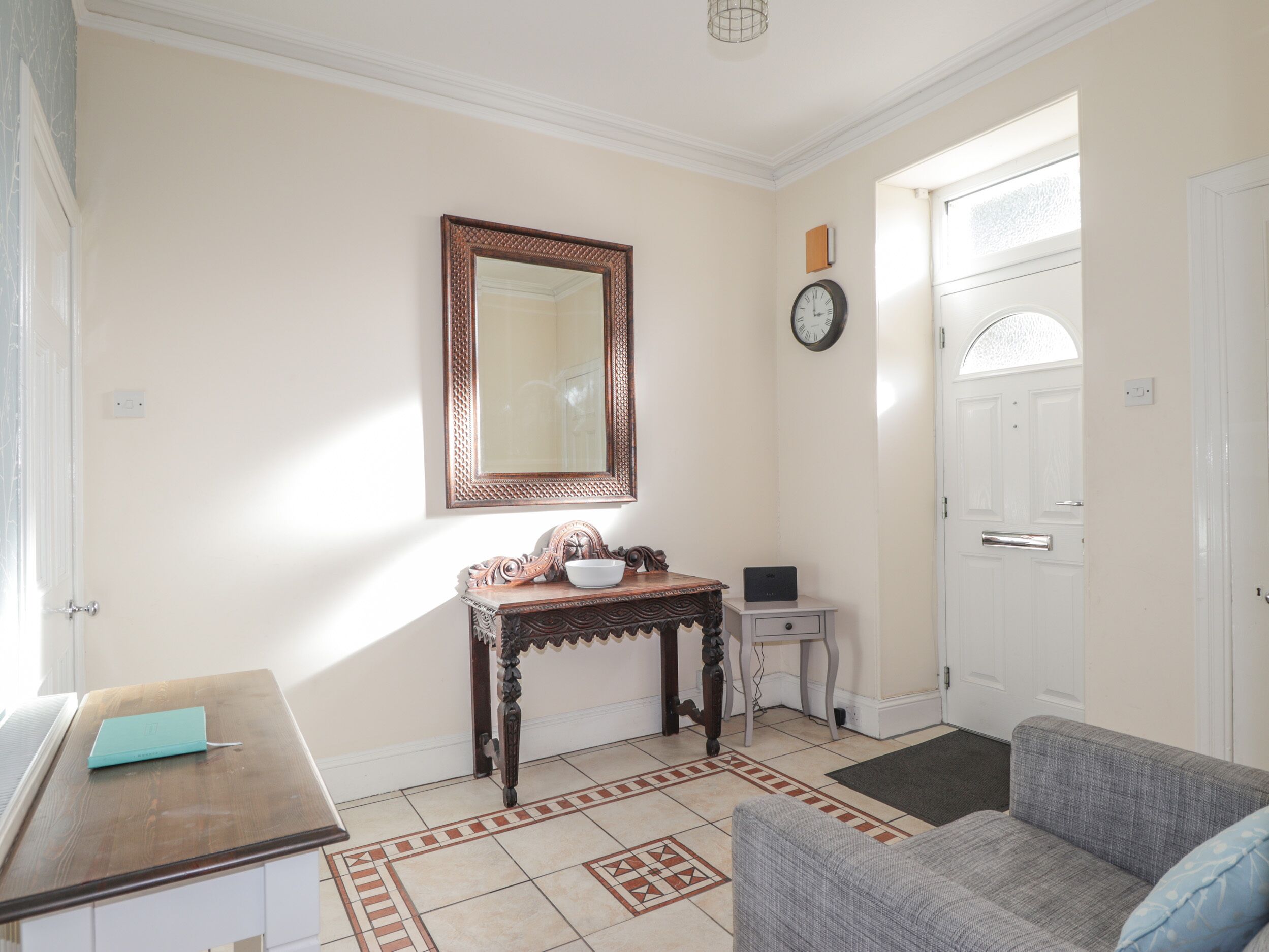 A room with a wooden console table, a white chair, a grey sofa, a large mirror, and a clock on the wall.