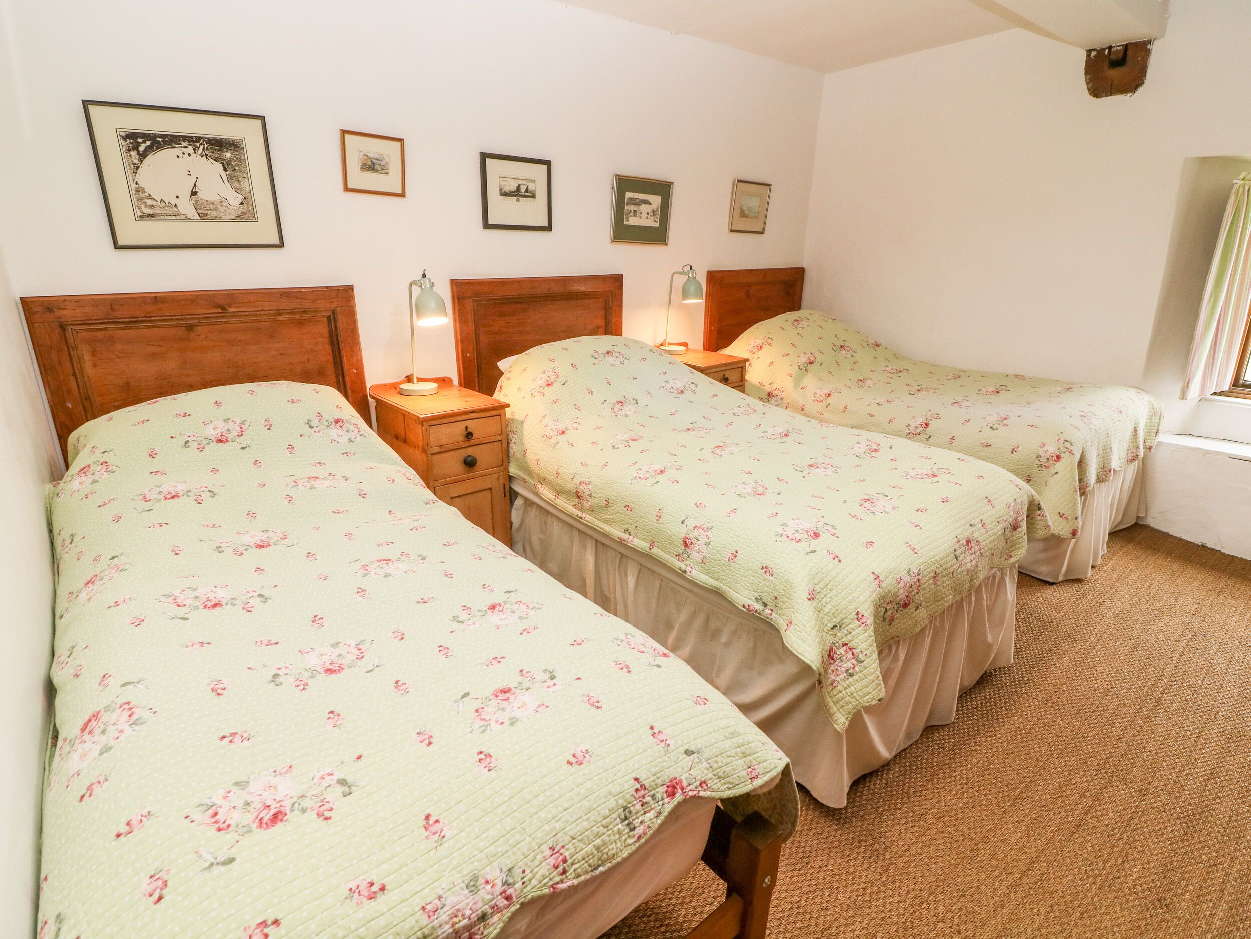 A room with two beds, each with floral bedding and wooden headboards. There are framed pictures on the wall and a small bedside table with a lamp.