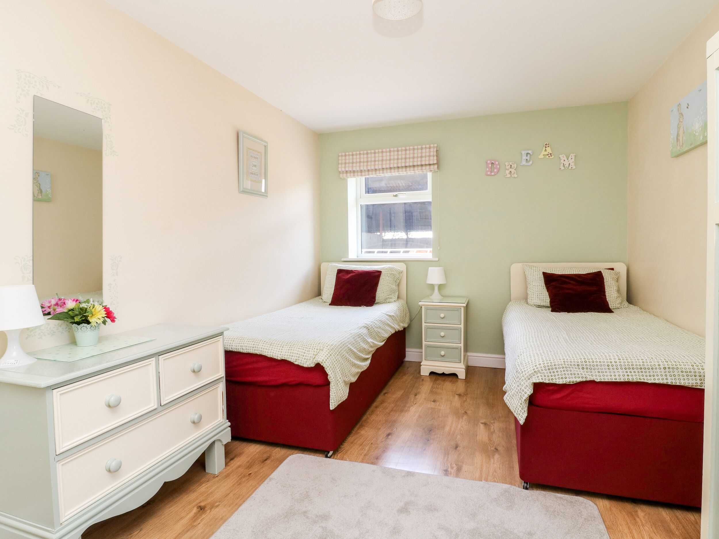 A bedroom with two beds, a dresser, a nightstand, a window with a checkered valance, and a wall with the word "DREAM" spelled out.