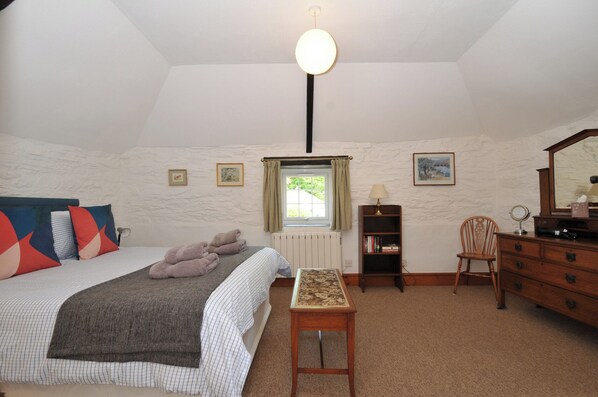 A bedroom with a bed, a bedside table, a dresser, a mirror, a window with curtains, and a wall-mounted light fixture. - Trevenning Barn (Bodmin)