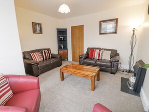 A living room with two brown leather sofas, a wooden coffee table, and a red armchair. There is a blue cabinet with shelves, a wooden door, and a floor lamp. - Gwelfor (Porthmadog)