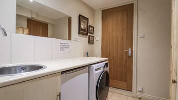 A compact kitchen with a sink, a washing machine, and a wooden door. A sign requests placing recyclable glass and plastics in a box.