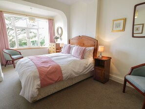 Un dormitorio ordenado con una cama grande, una silla, un aparador y una mesita de noche con lámpara.