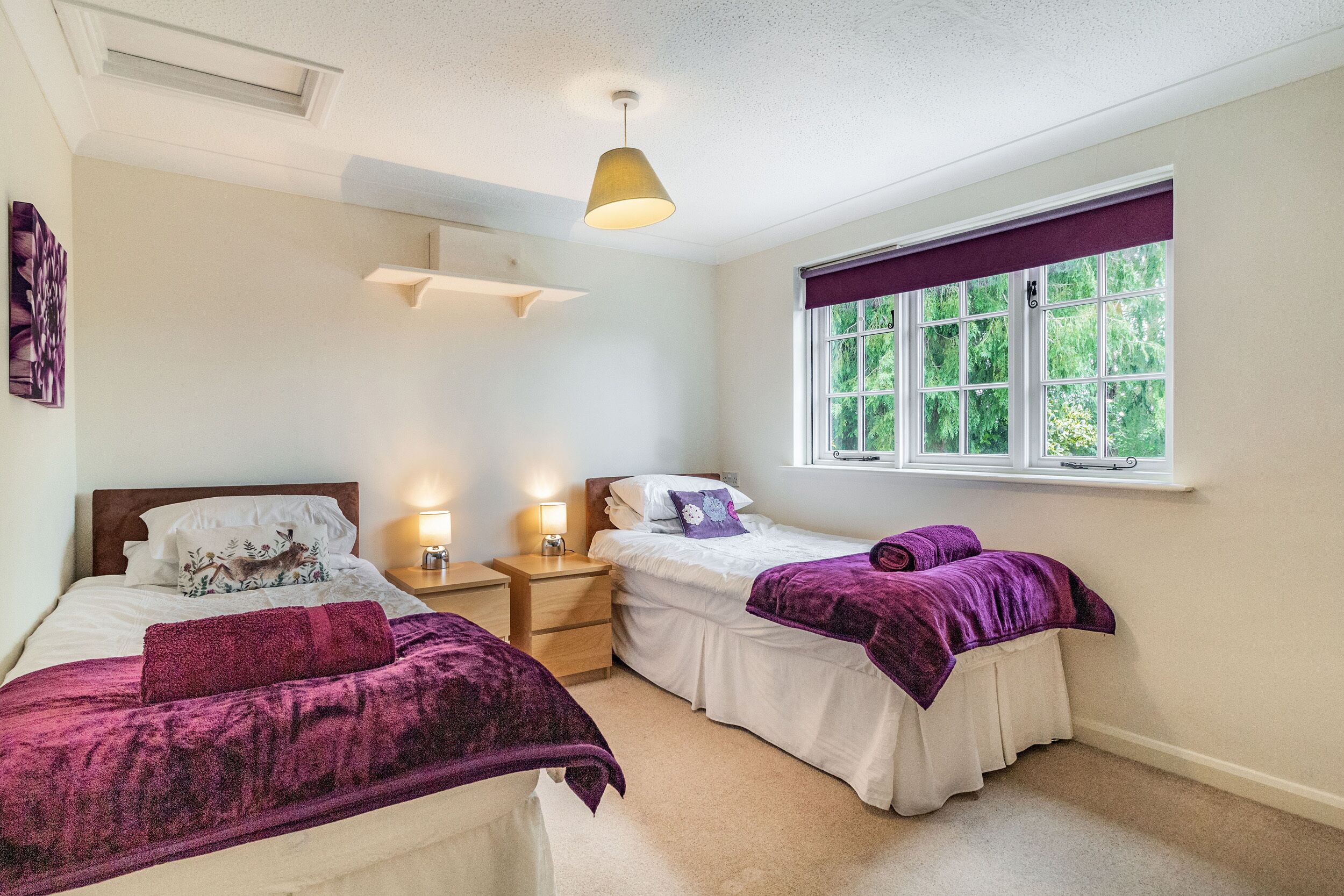 A bedroom with two beds, a window with a view of greenery, a ceiling light, and a wall-mounted shelf.