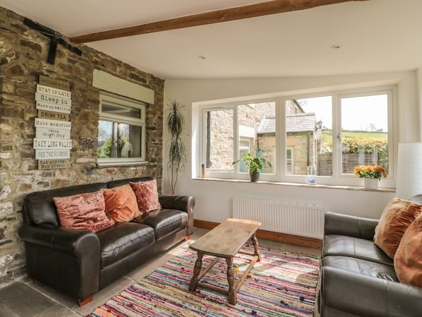 Un salon chaleureux avec un mur en pierre, un canapé en cuir noir, une table basse en bois et une vue sur la campagne, visible à travers de grandes fenêtres.