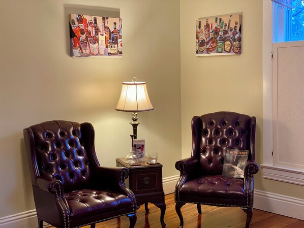 Two leather armchairs, a small side table with a lamp, and two wall-mounted paintings of liquor bottles. - Red Rose Inn Bed & Breakfast (Bardstown)
