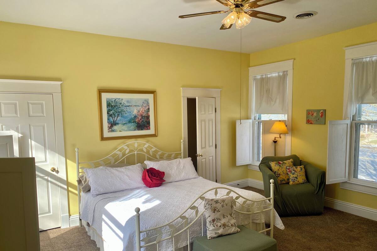A bedroom with a bed, a green chair, a painting, and a ceiling fan.