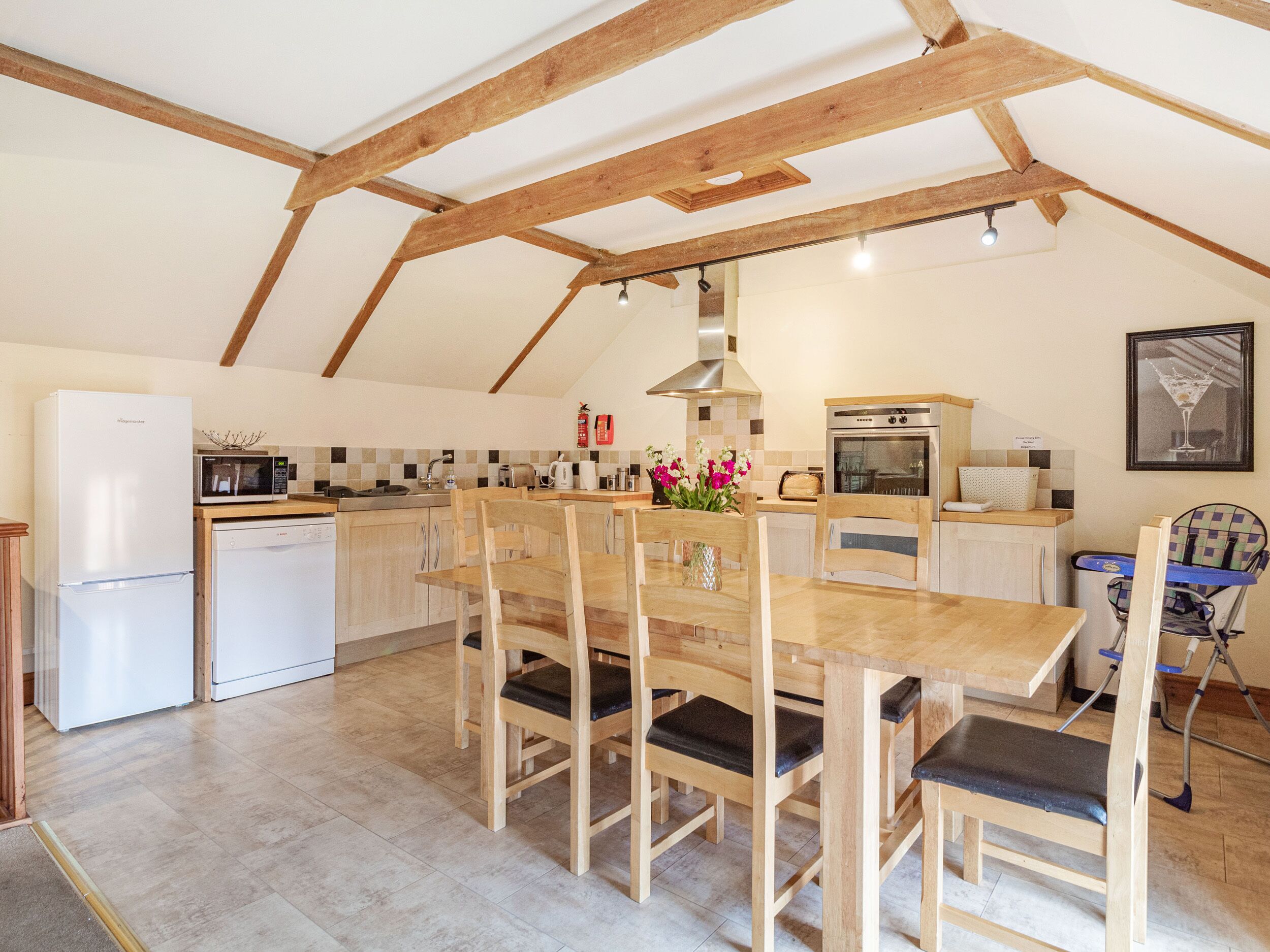Een keuken met houten balken, een eettafel met stoelen, een koelkast, een magnetron, een fornuis en een spoelbak.