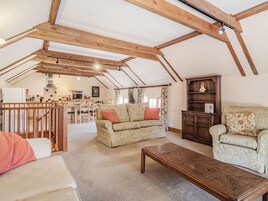 Un loft espacioso con vigas de madera a la vista, una zona de estar acogedora con sofá y sillón, una mesa de centro de madera, y una cocina al fondo.