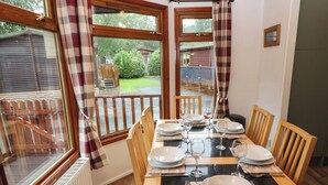 A dining area with a table set for four, large windows with curtains, and a view of a wooden deck and greenery outside. - Langdale Lodge 15 (Windermere)