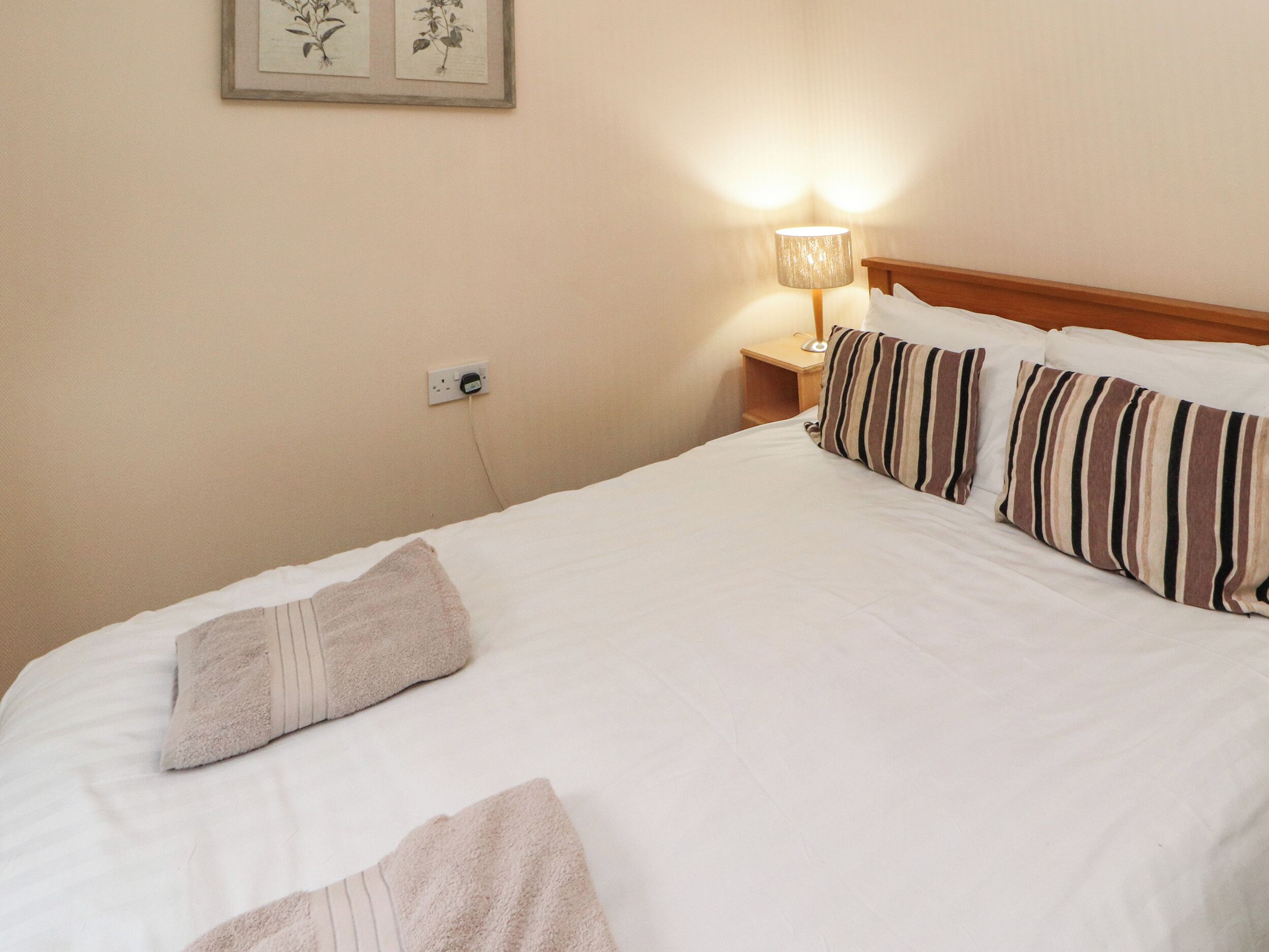 A neatly made bed with striped pillows and a white blanket, a bedside lamp, and a wall-mounted power outlet.