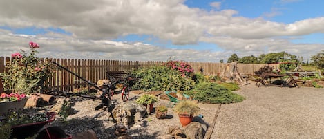 Une allée de gravier mène à une clôture en bois, agrémentée de plantes en pot et d’une brouette.