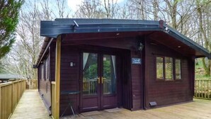A wooden cabin with a covered porch, a small table, and a sign indicating the name. - Top Lodge, 4 Skiptory Howe (Windermere)