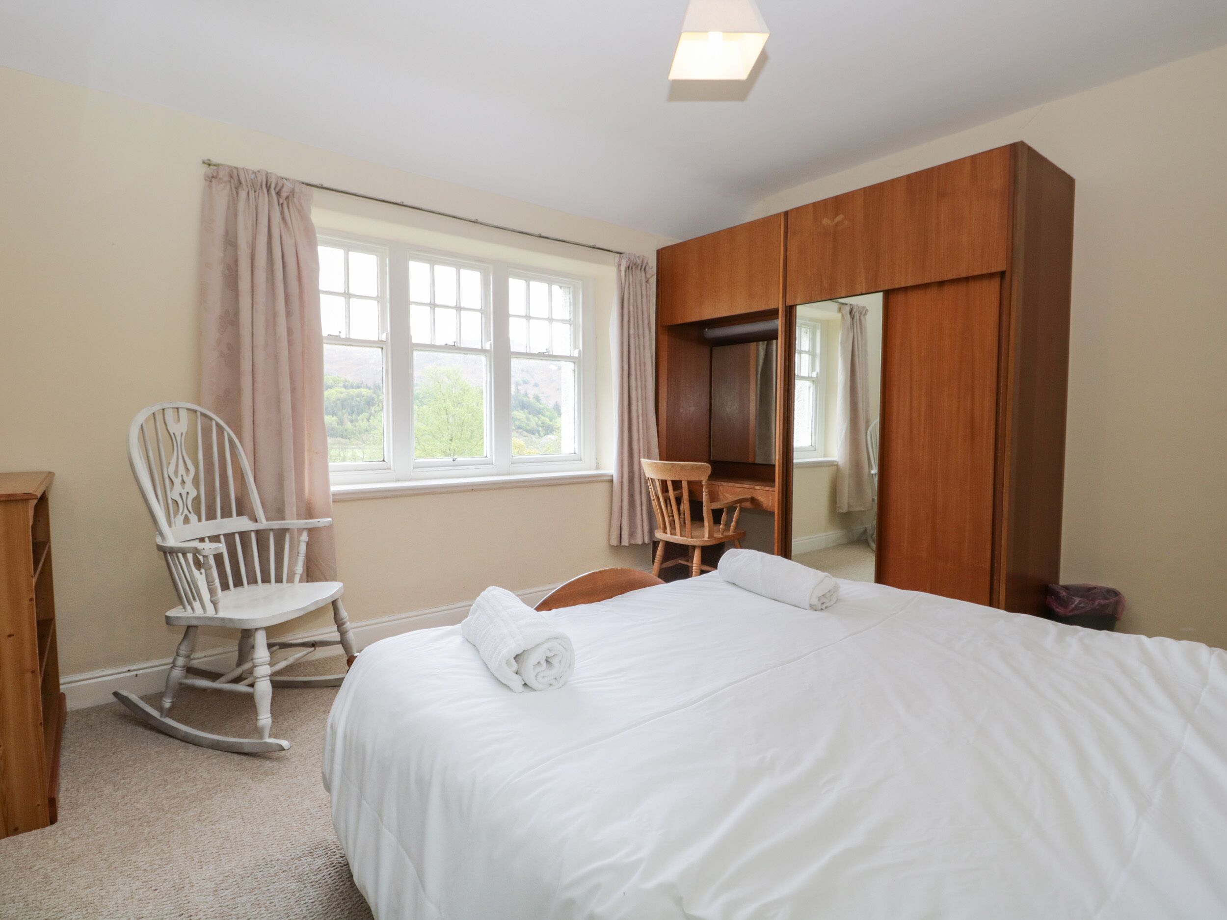 A bedroom with a bed, two rocking chairs, a wooden wardrobe, and a window with curtains.