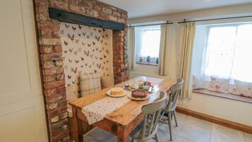 Un coin repas avec un mur en briques, une table en bois et des chaises. Il y a une fenêtre avec des rideaux et une vue sur l’extérieur.
