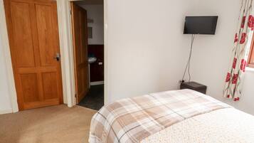 A bedroom with a wooden door, a bed with a plaid bedspread, a television on the wall, and a bathroom visible through an open door.