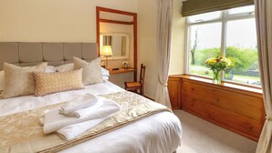 A neatly made bed with pillows and folded towels, a bedside table with a lamp, a chair, and a window with curtains. - Longwell House (Kendal)