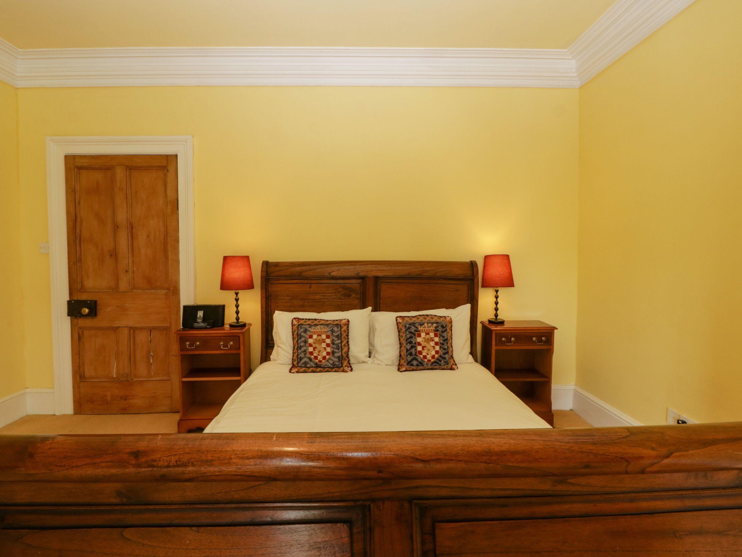 Un dormitorio con una cama de madera, dos mesitas de noche con lámparas, una puerta de madera y paredes amarillas.