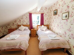 A bedroom with two single beds, a wooden nightstand, a window with curtains, and floral wallpaper. - Barnbrook (Leyburn)