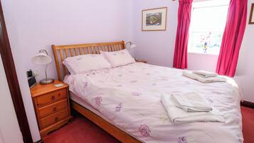 A neatly made bed with floral bedding, a bedside lamp, and a window with pink curtains.