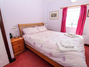 Una cama bien hecha con ropa de cama floral, una lámpara de noche y una ventana con cortinas rosadas.