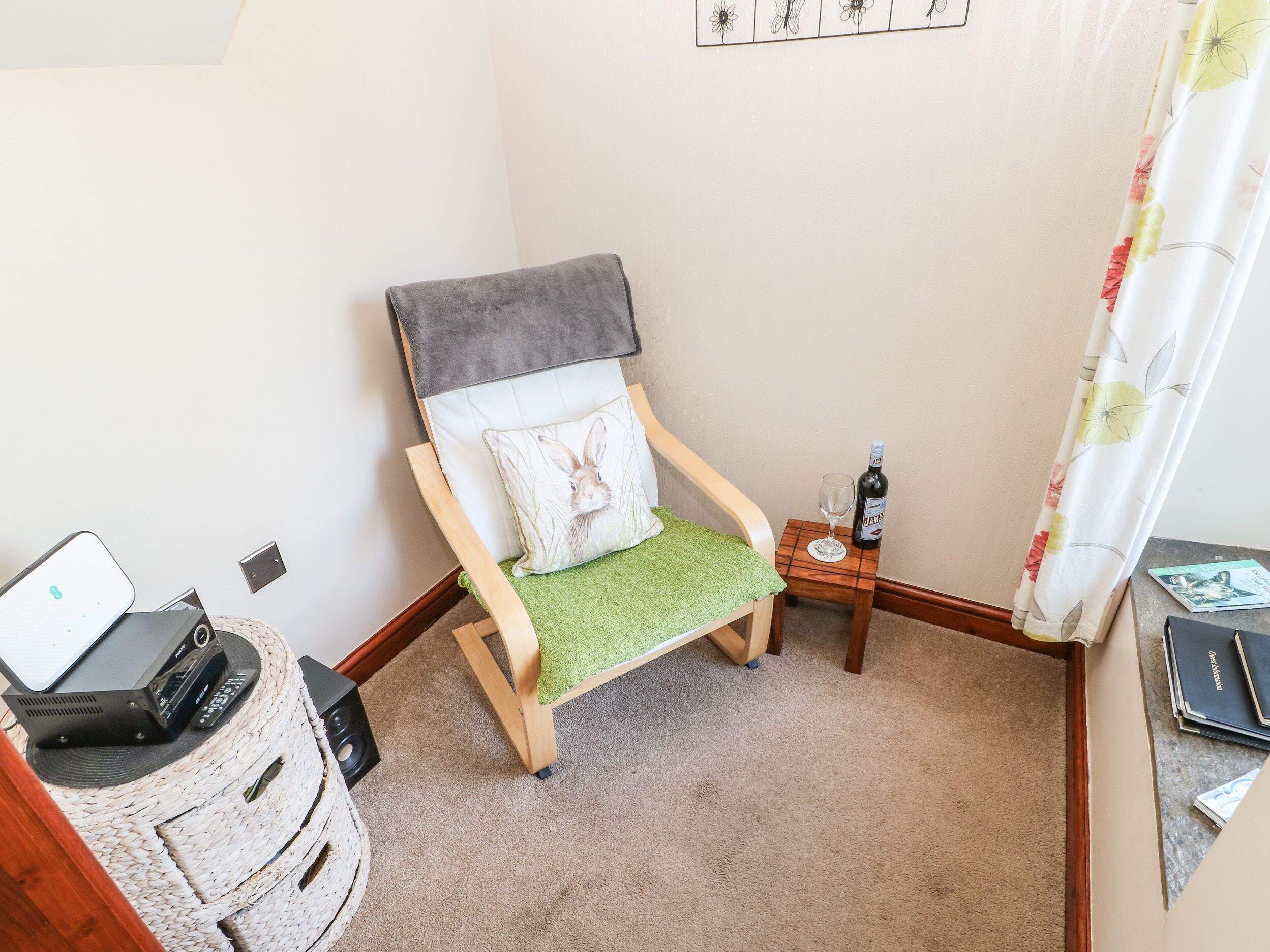 Een hoekzaal met een houten stoel met een groene kussen, een klein tafeltje met een fles en glas, en een laptop op een rieten mand.