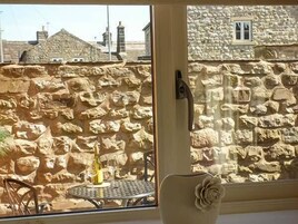 Quarto com parede de pedra, janela com vista para casas vizinhas, cadeira com detalhe floral e uma garrafa sobre uma mesa.