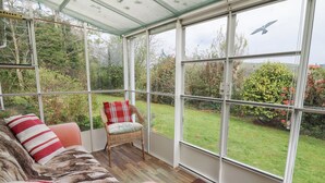 A conservatory with a wicker sofa, a chair, and a view of a garden. - Bryn yr Haf (Lampeter)