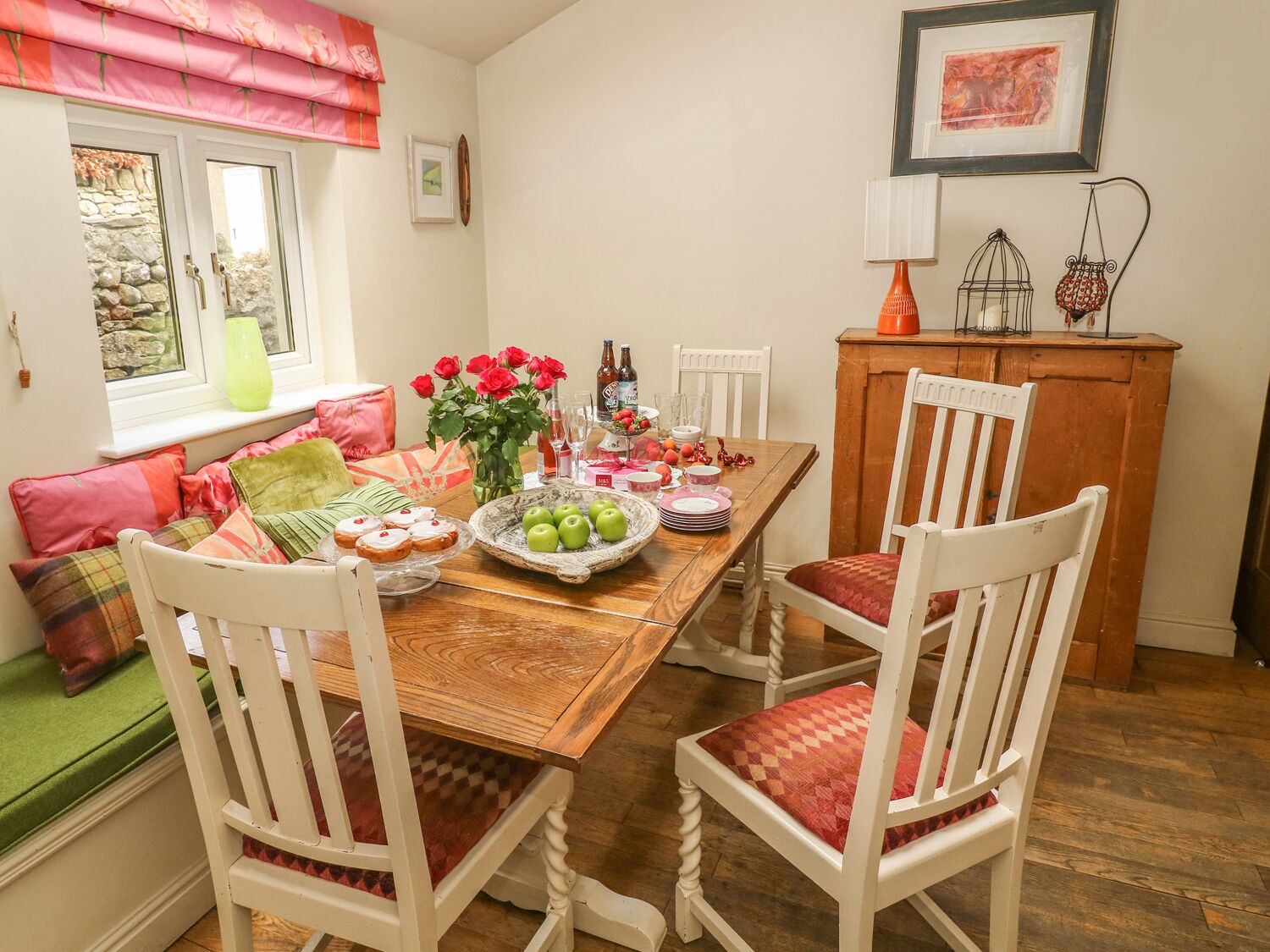 A dining area with a wooden table set for four, surrounded by white chairs with red cushions. A vase with red roses and a fruit bowl are on the table.