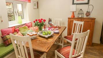 A dining area with a wooden table set for four, surrounded by white chairs with red cushions. A vase with red roses and a fruit bowl are on the table.