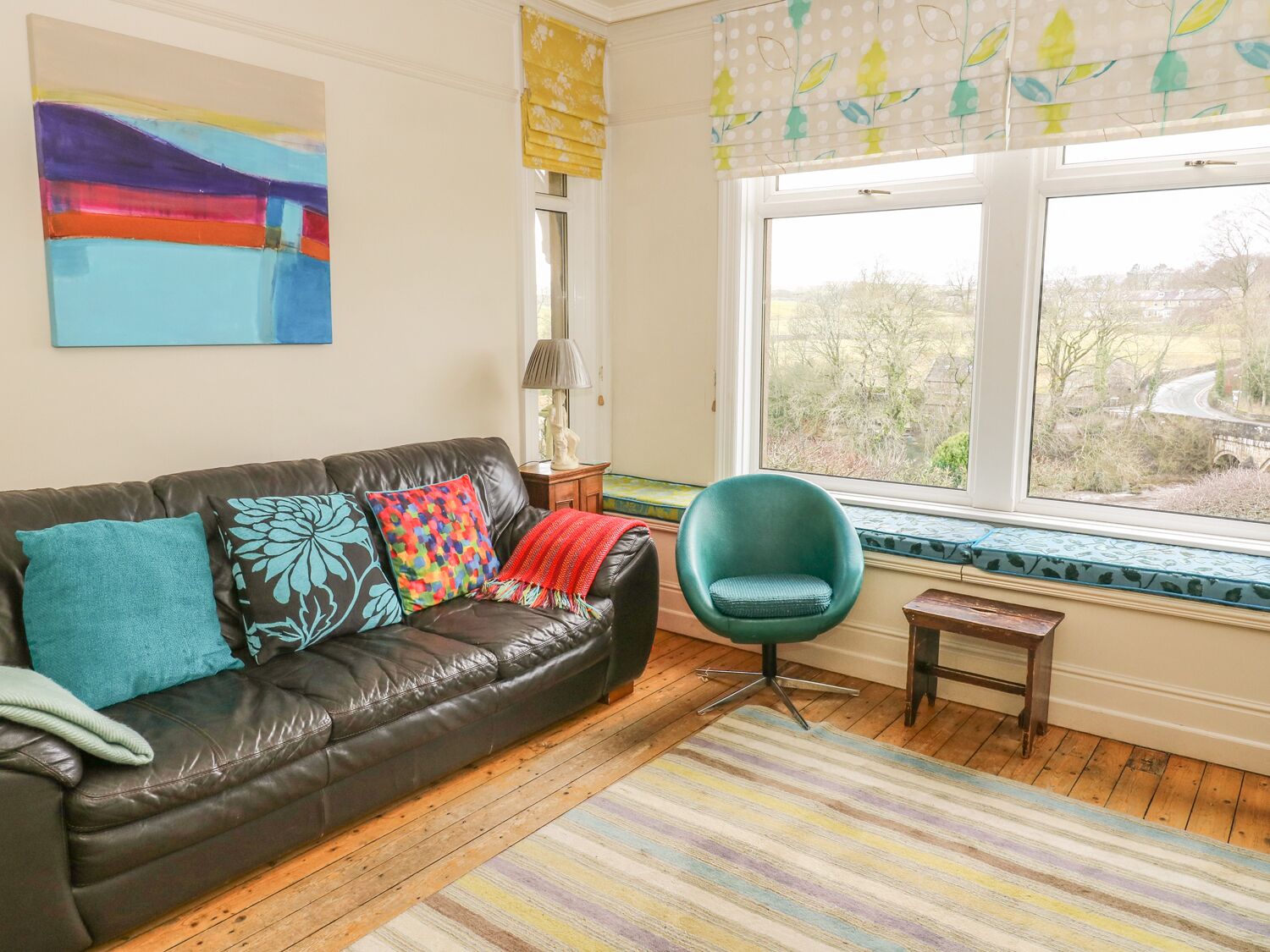 A living room with a leather sofa, colorful cushions, a teal armchair, a small wooden table, and a view of a bridge through a window with a leaf-patterned valance.